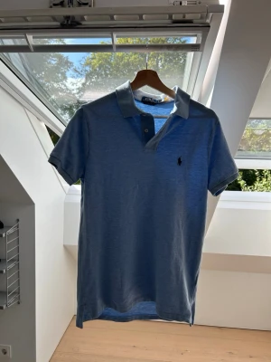 Blå pikétröja från Ralph Lauren - Snygg blå pikétröja från Ralph Lauren med klassisk krage och korta ärmar. Tröjan har den ikoniska loggan broderad på bröstet och är tillverkad i mjuk bomull. Perfekt till jeans eller shorts för en avslappnad stil.