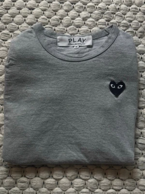 Grå t-shirt från Comme des Garçons  - Grå t-shirt från Comme des Garçons Play med det ikoniska svarta hjärtat med ögon broderat på bröstet. Klassisk rund halsringning och korta ärmar. Tillverkad i mjuk bomull för en skön och avslappnad stil.