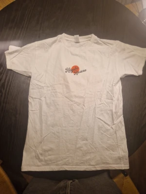 Vit t-shirt med Högsbo Riviera-tryck - Säljer en vit t-shirt med trycket 'Högsbo Riviera' i retrostil. Framsidan har en liten logga medan baksidan har ett stort tryck med sol, vågor och en badande person i orange och svart. T-shirten har rund hals och korta ärmar, perfekt för en avslappnad look.