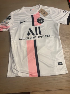 PSG matchtröja Messi nr 30 - Snygg vit PSG matchtröja med rosa och svart rand framtill, Messi och nummer 30 på ryggen. Tröjan har officiella klubbmärket, Champions League-märke på ärmen och sponsortryck. Tillverkad i lätt och ventilerande material, perfekt för fotboll eller träning.