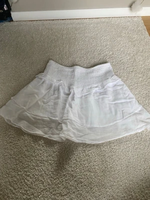 Vit kortkjol från Gina Tricot - Säljer en vit kortkjol från Gina Tricot med bred smock i midjan och lätt, luftig känsla. Kjolen har en volangig nederdel och är perfekt för varma dagar. Tillverkad i ett tunt och mjukt material som ger en skön passform.
