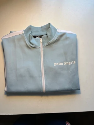 Ljusblå ziptröja från Palm Angels - Snygg ljusblå ziptröja från Palm Angels med vit dragkedja och vita ränder längs ärmarna. Tröjan har ribbade muddar, ståkrage och fickor med dragkedja. Perfekt för en sportig och trendig look.