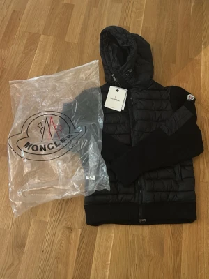 Moncler cardigan - Snygg svart pufferjacka från Moncler med huva och dragkedja framtill. Jackan har quiltad kropp i glansigt material och stickade ärmar för en modern look. Moncler-logga på vänster ärm och ribbade muddar vid ärmslut och nederkant. Perfekt för kalla vinterdagar.
