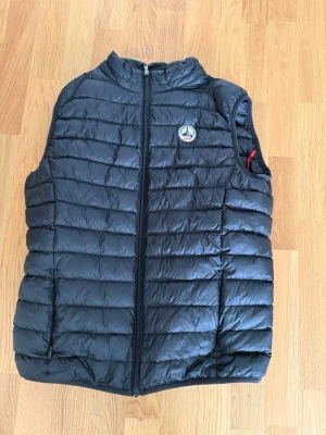 Svart dunväst från Jott  - Snygg svart dunväst med horisontella sömmar och ståkrage. Västen har dragkedja framtill och en liten rund logotyp på bröstet. Perfekt för lager-på-lager och enkel att matcha med olika outfits.