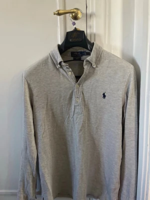 Ljusgrå långärmad henley pikétröja från Polo Ralph Lauren - Säljer en stilren ljusgrå långärmad henley från Polo Ralph Lauren. Skjortan har klassisk krage, knappar framtill och den ikoniska mörkblå loggan broderad på bröstet. Tillverkad i mjuk bomull som känns lätt och bekväm. Perfekt för en clean och avslappnad look.