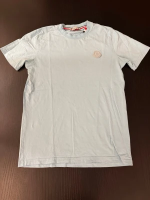 Ljusblå t-shirt moncler - Snygg ljusblå t-shirt i bomull med rund hals och korta ärmar. På bröstet sitter en diskret patch som detalj. Perfekt för en clean och enkel stil.