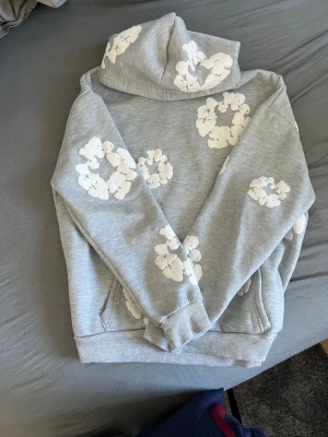  Denim tears Grå hoodie med vita blommor  - Snygg grå hoodie med huva och vita blommönster tryckta över hela tröjan. Hoodien har en klassisk passform, ribbade muddar och en stor ficka framtill. Perfekt för dig som vill sticka ut med en unik och trendig stil.