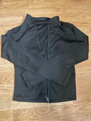 Svart sportjacka med dragkedja - Snygg svart sportjacka med hel dragkedja framtill och hög krage. Tillverkad i ett stretchigt och lätt material som passar perfekt för träning eller löpning. Jackan har långa ärmar och två sidofickor. Enkel och stilren design som funkar till det mesta.