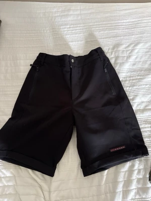 Prada Kostymshorts – Svarta - Prada Kostymshorts – Svarta 🩳 Typ: Kostymshorts / dressade shorts 🖤 Färg: Svart ✨ Skick: 7/10 – använda men fortfarande i bra skick 🔒 Detaljer: Fickor med dragkedja framtill 🔺 Design: Prada