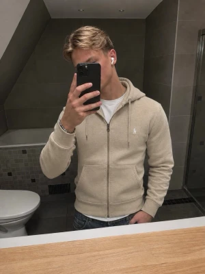 Beige hoodie från Polo Ralph Lauren - Snygg beige hoodie från Polo Ralph Lauren med dragkedja framtill och klassisk huva. Tröjan har två fickor och den ikoniska lilla loggan broderad på bröstet. Perfekt till jeans eller sweatpants för en clean och stilren look.