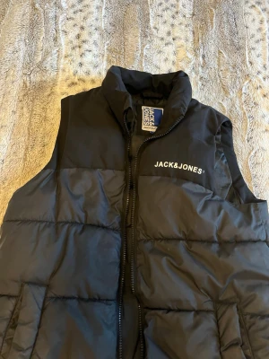 Svart dunväst från Jack & Jones - Snygg svart dunväst från Jack & Jones med hög krage och dragkedja framtill. Västen har broderad logga på bröstet och är vadderad för extra värme. Perfekt att slänga över en hoodie för en cool look.