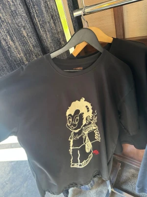 House of savojxLugazi svart t shirt  - Cool svart t-shirt med ett stort grafiskt tryck framtill som föreställer en tecknad figur med glasögon och en kopp. T-shirten har rund hals och korta ärmar. Perfekt för dig som gillar streetwear och unika prints.
