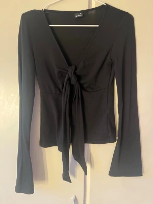 Svart långärmad topp med knyt - Trendig svart långärmad topp med djup v-ringning och knyt framtill. Toppen har en figurnära passform och är tillverkad i ett mjukt, stretchigt material som ger en bekväm känsla. Perfekt att styla med jeans eller kjol för en cool look.