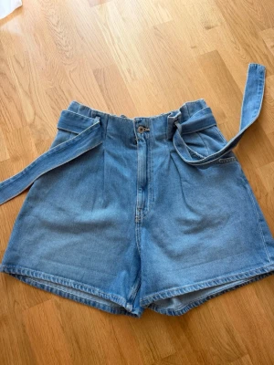 Högmidjade jeansshorts med bälte - Säljer ett par ljusblå högmidjade jeansshorts med tillhörande bälte. Shortsen har en relaxed passform, knapp- och dragkedjestängning samt fickor både fram och bak. Perfekta för varma dagar och enkel att styla med olika toppar.