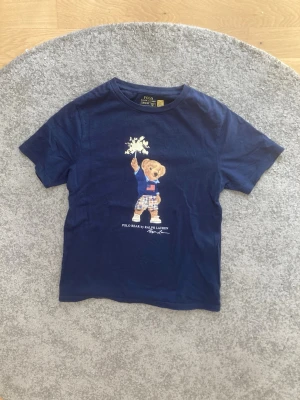 Marinblå Polo Bear t-shirt Ralph Lauren - Säljer en marinblå t-shirt från Ralph Lauren med den ikoniska Polo Bear-figuren på bröstet. T-shirten är kortärmad och har en klassisk passform. Tillverkad i mjuk bomull som känns skön mot huden. Perfekt för dig som gillar stilrena och lekfulla plagg.
