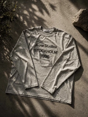 Acne studios long sleeve - Snygg grå långärmad topp från Acne Studios med tryck 'STOCKHOLM 1996' framtill. Tillverkad i mjuk bomull och har en avslappnad passform. Perfekt för en clean och stilren look.