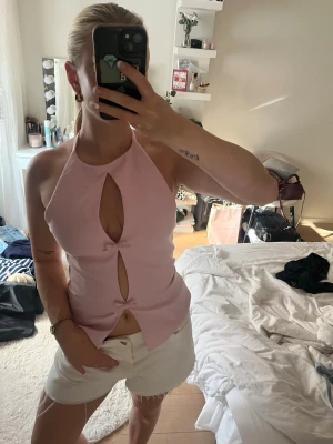 Ljusrosa halterneck topp med rosetter - Säljer en ljusrosa halterneck topp med två dekorativa rosetter framtill och snygga cut-outs. Toppen är ärmlös och har en figurnära passform som ger en trendig look. Perfekt för dig som vill sticka ut lite extra i sommar.