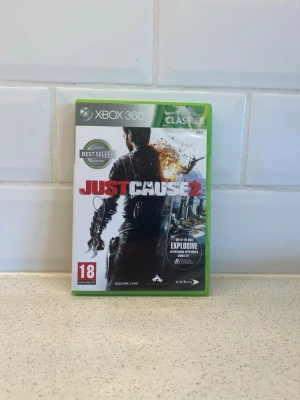 Just Cause 2 till Xbox 360 - Just Cause 2 är ett actionspel till Xbox 360 med öppen värld och massor av explosioner. Omslaget är grönt med en cool illustration av huvudkaraktären och stadsmiljö. Spelet är märkt 18+ och har engelska texter. Perfekt för dig som gillar adrenalinfyllda äventyr. Fungerar toppen!