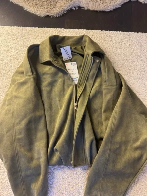 Olivgrön mockajacka med dragkedja - Snygg olivgrön jacka i mockaimitation med dragkedja framtill och klassisk krage. Jackan har en stilren design och passar perfekt till många olika outfits. Materialet ger en mjuk känsla och jackan har en modern passform.