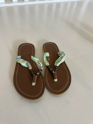 Bruna sandaler med färgglada band - Säljer ett par bruna sandaler med mjuk sula och färgglada tygband i pastellfärger. Remmarna är dekorerade med pärlor och har en bohemisk känsla. Perfekta för sommaren och strandhäng. Snygg kontrastsöm runt sulan och blommönster på fotbädden.