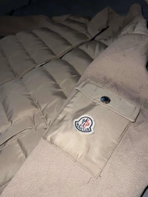 Beige dunjacka  - Säljer en beige dunjacka med klassisk quiltad design och dragkedja framtill. Jackan har en ficka med knapp på ärmen och Insidan är vit med en serietidningsdetalj och innerficka. Perfekt för kalla vinterdagar.