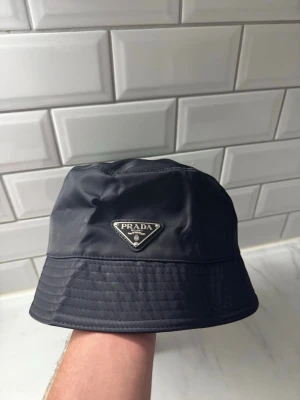 Svart bucket hat från Prada - Snygg svart bucket hat från Prada med klassisk trekantig logotyp framtill. Hatten är tillverkad i ett glansigt nylonmaterial som ger en modern och stilren look. Perfekt accessoar för att lyfta din outfit med en exklusiv touch.