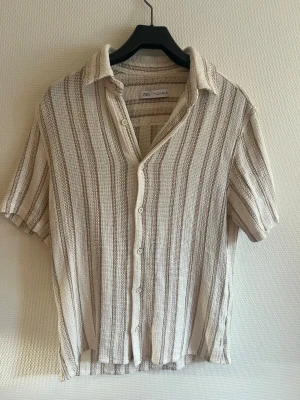 Randig skjorta från Zara i bomull - Säljer en luftig och avslappnad skjorta från Zara i beige och vit med vertikala ränder. Skjortan har korta ärmar, knappar framtill och är tillverkad i ett mjukt bomullstyg med strukturerad känsla. Perfekt för varma dagar!