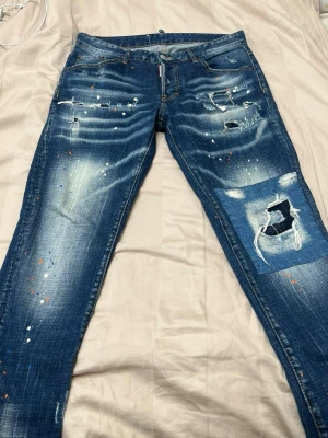 Blå distressed jeans från Dsquared2 - Säljer ett par blå jeans från Dsquared2 med slitningar, hål och patchdetaljer. Jeansen har skinny fit, orange kontrastsömmar och färgstänk för en edgy look. Perfekta för dig som gillar streetwear och vill sticka ut. Storlek 48 i dsquared jeans är som 32 i vanliga!