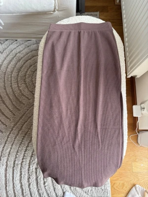 Ribbad kjol - Säljer en rosa/lila ribbad kjol i mjukt stickat material. Kjolen har en rak passform och elastisk midja för extra komfort. Perfekt att styla med en enkel topp eller stickad tröja. Har en hög slits.