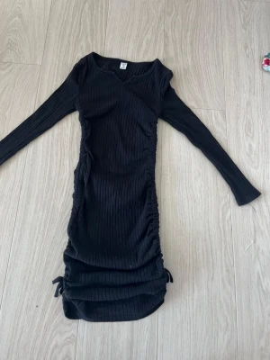 Svart ribbad klänning  - Snygg svart ribbad klänning med omlottdetalj och långa ärmar. Klänningen har snörning i sidorna och är i kort modell. Perfekt för dig som gillar en tight och trendig look.