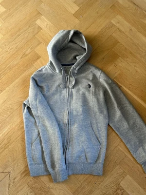 Grå Ralph lauren zip hoodie  - Snygg grå hoodie med dragkedja och huva. Klassisk passform med två fickor framtill och justerbara snören i huvan. Liten broderad logga på bröstet. Tillverkad i mjukt sweatshirtmaterial som är skönt mot huden. Märket är U.S.POLO ASSN. Finns även likande plagg i profilen. Storlek S men passar typ som M