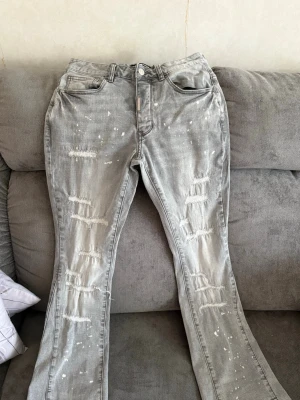 Grå slitna jeans från Lorenzo - Säljer ett par grå jeans från Lorenzo med slitningar och hål på benen samt vita färgstänk för en trendig look. Jeansen har klassisk femficksmodell och lätt utsvängda ben. Perfekta för dig som gillar streetstyle och vill sticka ut. UK size 28 motsvarar W29 L30