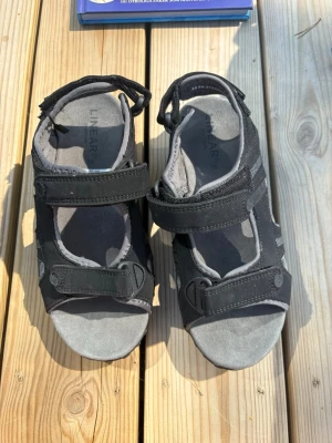 Svarta sportiga sandaler med kardborre - Säljer ett par svarta sandaler med grå detaljer och justerbara kardborreband. De har en sportig design med öppna sidor och slitstark sula, perfekta för sommarens äventyr. Märket Linear syns på innersulan. Materialet är en mix av textil och syntet.