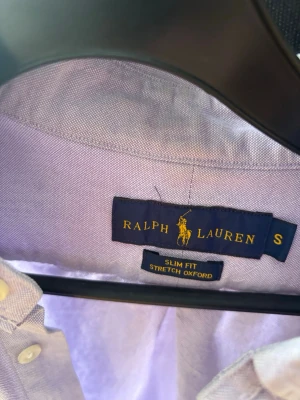 Ljuslila skjorta från Ralph Lauren - Säljer en ljuslila slim fit skjorta från Ralph Lauren i stretchig oxford-bomull. Skjortan har klassisk krage, knappar framtill och den ikoniska broderade loggan på bröstet. Perfekt för dig som gillar stilrena och tidlösa plagg.
