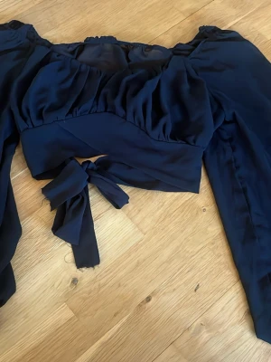 Svart omlottopp med knyt - Trendig svart omlottopp med långa ärmar och snygg knytning framtill. Toppen har en croppad passform och är gjord i ett mjukt, lätt material som ger en luftig känsla. Perfekt för dig som vill ha en stilren men ändå unik look.