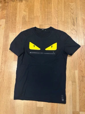 Fendi tshirt - Cool svart t-shirt med tryck av två gula ögon på bröstet och en dekorativ dragkedja under ögonen. T-shirten har rund hals och korta ärmar. Perfekt för dig som vill sticka ut med en unik och edgy stil.