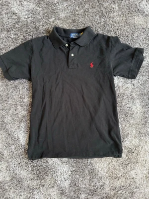 Svart pikétröja från Polo Ralph Lauren - Klassisk svart pikétröja från Polo Ralph Lauren med korta ärmar och krage. Tröjan har två knappar framtill och den ikoniska röda logotypen broderad på bröstet. Perfekt till jeans eller shorts för en clean look.