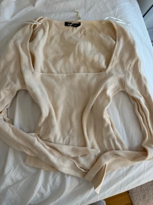 Beige ribbad långärmad topp - Trendig beige ribbad topp med lång ärm och fyrkantig urringning. Tillverkad i mjuk bomullsblandning och har en figurnära passform som framhäver siluetten. Perfekt att styla med jeans eller kjol för en clean look.