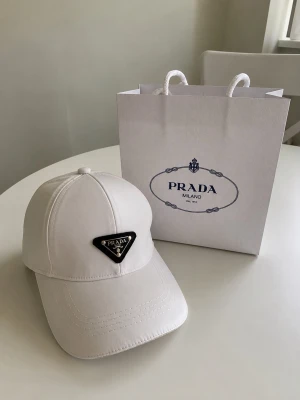 Vit keps från Prada - Snygg vit keps från Prada med klassisk böjd skärm och ikonisk svart metallogo framtill. Tillverkad i ett glansigt material som ger en exklusiv känsla. Perfekt accessoar för dig som vill ha en stilren och trendig look. Påsen kostar 100kr extra om man vill ha med den :) inköpt 2023 på rea för 3500 i Milano. 