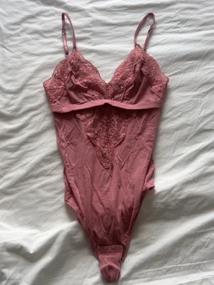 Rosa spetsbody med smala axelband - Säljer en snygg rosa body med spetsdetaljer och smala justerbara axelband. Bodyn har v-ringning, spets över bysten och ett dekorativt spetsmönster framtill. Materialet är mjukt och stretchigt, perfekt för en trendig look.