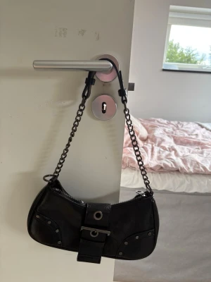 Svart axelremsväska med kedja - Snygg svart handväska i skinnimitation med cool kedjerem och detaljer som nitar och bälte framtill. Väskan har en böjd form och dragkedja upptill. Perfekt accessoar för att lyfta din outfit.