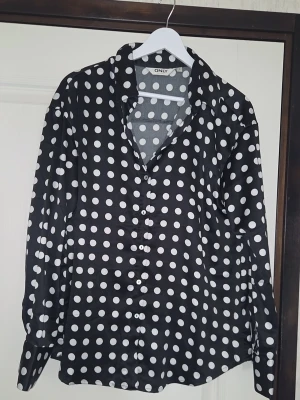 Svart prickig blus från ONLY - Snygg svart blus från ONLY med vita stora prickar och knäppning framtill. Blusen har lång ärm och klassisk krage, perfekt för en trendig och lekfull look. Materialet känns lätt och följsamt mot kroppen.