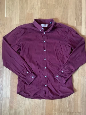 Lindbergh - Elegant skjorta | Vinröd | Slim fit | Herr | Strl L - Säljer en vinröd skjorta från Lindbergh med klassisk krage och vita knappar. Skjortan har lång ärm och är tillverkad i mjuk bomull. Perfekt för dig som vill ha en stilren och enkel look. Mycket bra använt skick! Jag är öppen för bud😊