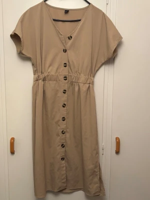Beige klänning med knappar och v-ringning - Stilren beige klänning med v-ringad hals och knappar längs hela framsidan. Klänningen har korta ärmar, resår i midjan och slits i sidan. Perfekt för en avslappnad och trendig look.