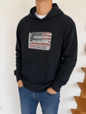 Svart hoodie från Ralph Lauren - Säljer en svart hoodie från Ralph Lauren med en stor amerikansk flagga och logga på bröstet. Hoodien har huva, magficka och ribbade muddar vid ärmar och nederkant. Perfekt för en avslappnad och cool stil.