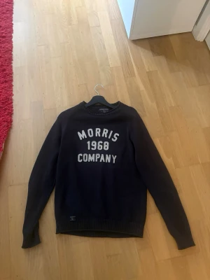 Mörkblå stickad tröja Morris - Mörkblå stickad tröja från Morris med vit text 'Morris 1968 Company' på bröstet. Tröjan har rund halsringning, långa ärmar och ribbade muddar vid ärmslut och nederkant. Perfekt för dig som gillar klassisk college-stil.