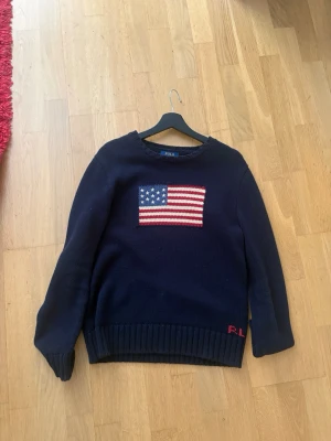 Mörkblå stickad tröja med USA-flagga - Mörkblå stickad tröja med en stor amerikansk flagga på bröstet. Tröjan har rund hals, långa ärmar och ribbstickade muddar vid ärmslut och nederkant. Perfekt för dig som gillar klassisk och stilren design.
