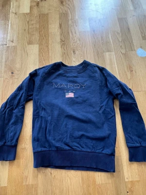 Mörkblå Marqy sweatshirt  - Mörkblå sweatshirt med rund hals och ribbade muddar.'MARQY' broderat ovanför en liten amerikansk flagga. Tröjan har en avslappnad passform och är gjord i mjukt bomullstyg.