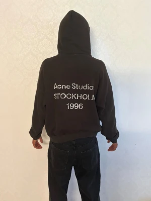 Svart hoodie från Acne Studios - Svart hoodie med huva från Acne Studios. På ryggen står det 'Acne Studios STOCKHOLM 1996' i vitt tryck. Hoodien har klassisk passform, ribbade muddar och är tillverkad i mjukt material. Perfekt för en avslappnad och trendig stil. Längd på personen på bild 182cm/Vikt 70kg
