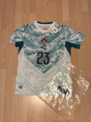 #23 Vitinha Portugal bortatröja Puma  - Snygg Portugal bortatröja från Puma med nummer 23 och Vitinha på ryggen. Tröjan har ett unikt turkost och vitt mönster, korta ärmar och rund hals. Perfekt för dig som älskar fotboll och vill stötta landslaget med stil.